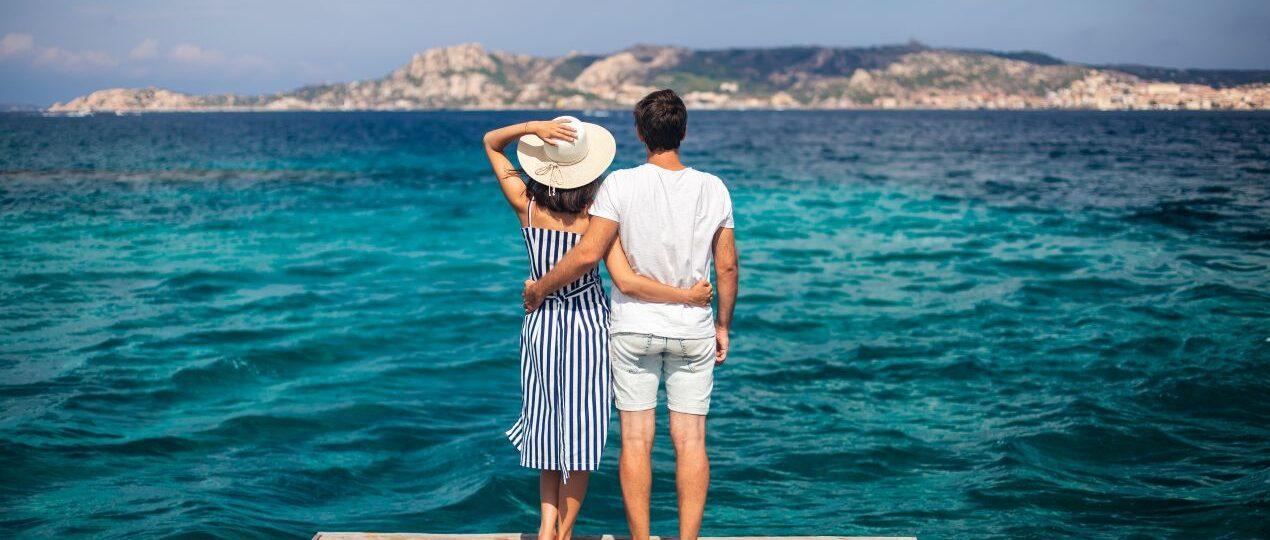 Ein Paar steht auf einem Holzsteg und blickt ueber das tuerkisfarbene Meer Richtung Kueste – idealer Moment fuer einen Urlaub im Sardinien Ferienhaus mit Hund.