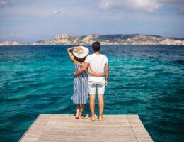 Ein Paar steht auf einem Holzsteg und blickt ueber das tuerkisfarbene Meer Richtung Kueste – idealer Moment fuer einen Urlaub im Sardinien Ferienhaus mit Hund.