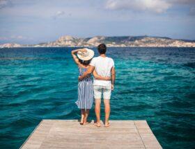 Ein Paar steht auf einem Holzsteg und blickt ueber das tuerkisfarbene Meer Richtung Kueste – idealer Moment fuer einen Urlaub im Sardinien Ferienhaus mit Hund.