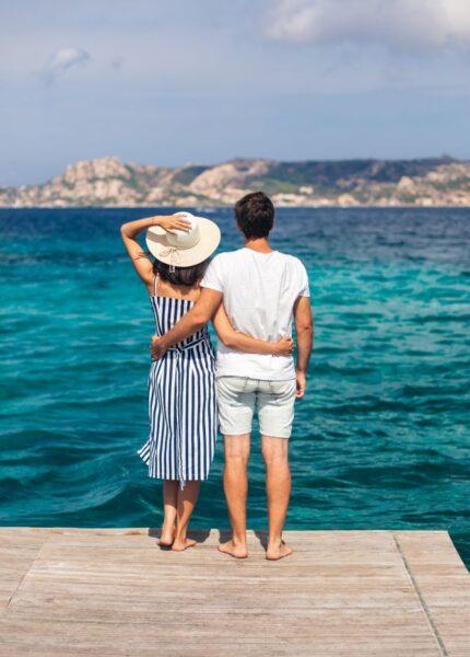 Ein Paar steht auf einem Holzsteg und blickt ueber das tuerkisfarbene Meer Richtung Kueste – idealer Moment fuer einen Urlaub im Sardinien Ferienhaus mit Hund.