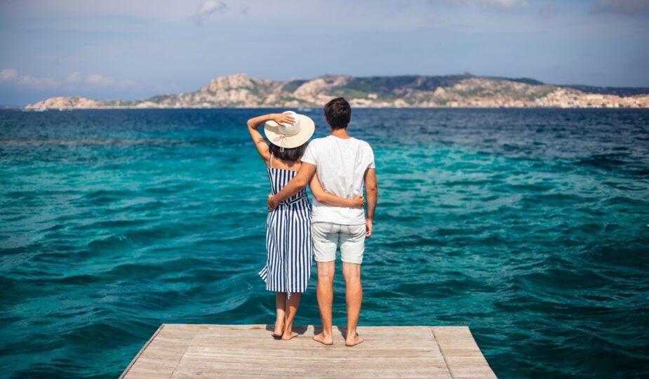 Ein Paar steht auf einem Holzsteg und blickt ueber das tuerkisfarbene Meer Richtung Kueste – idealer Moment fuer einen Urlaub im Sardinien Ferienhaus mit Hund.